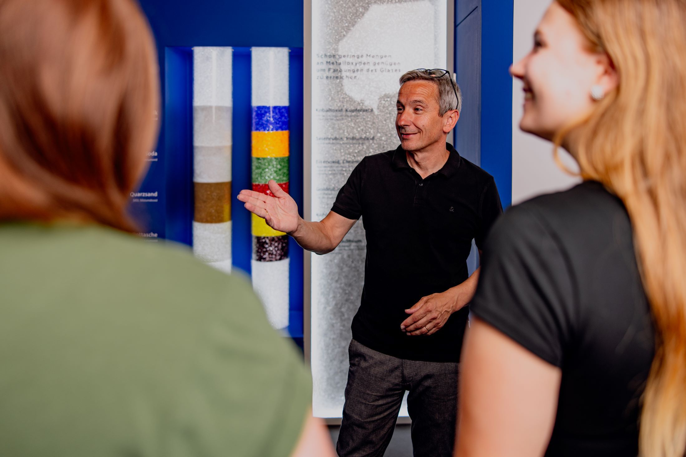 A man explains something to two women in an exhibition room with colorful glass tubes.