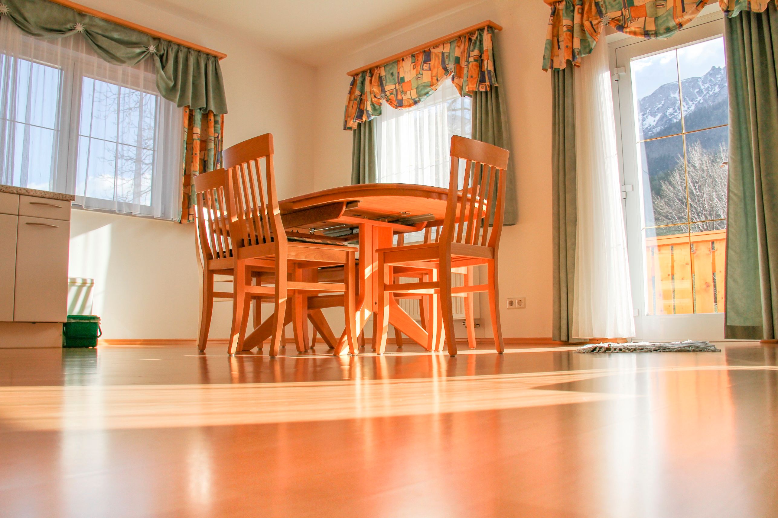 Dining area with wooden table and chairs in a sunny room with large windows and curtains.