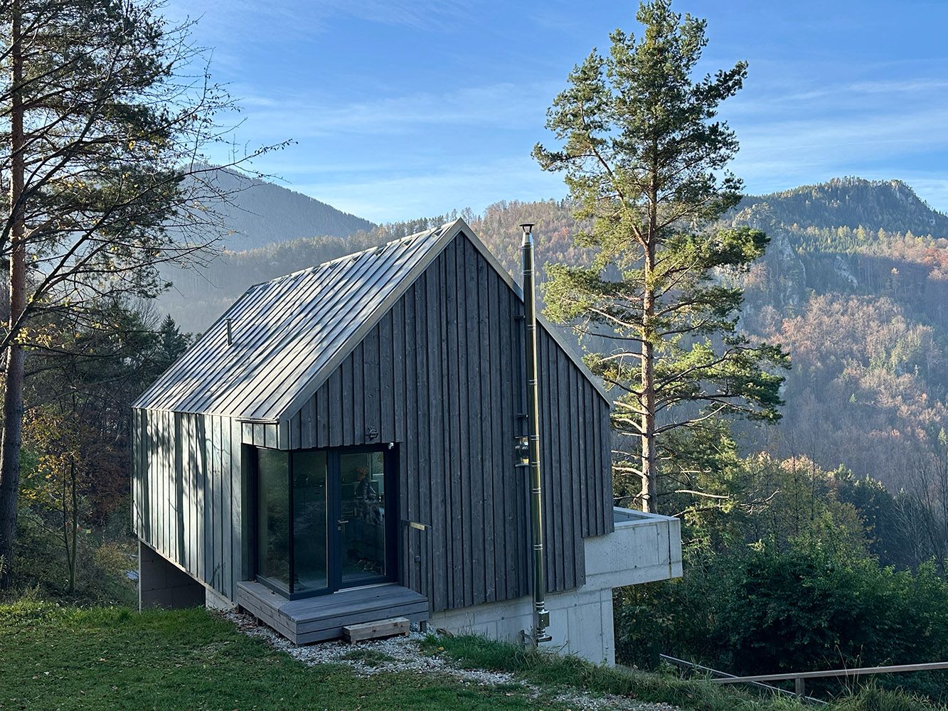 Modern wooden house in a mountainous landscape with trees and blue sky.