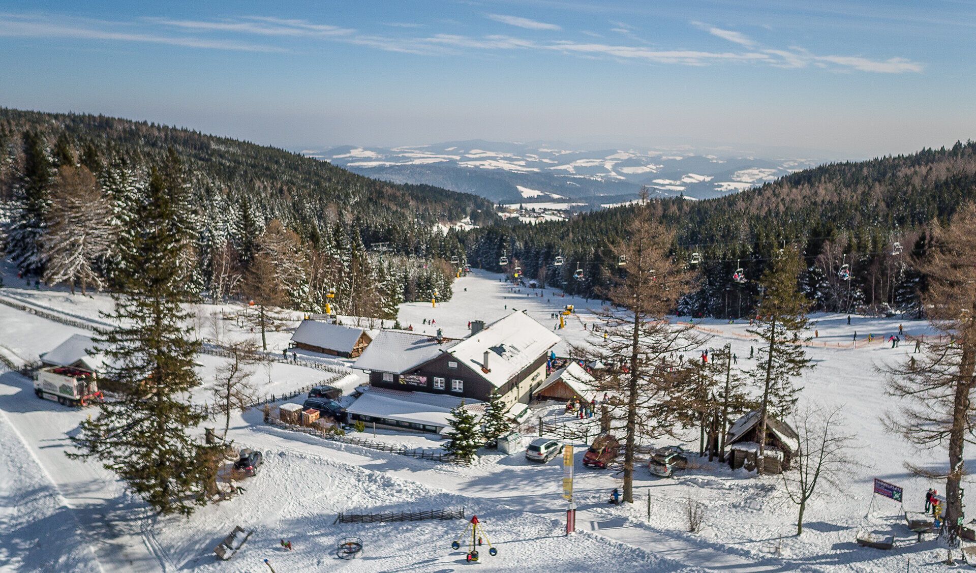 Ski area with chair lift and skiers, surrounded by snow-covered trees and hills.