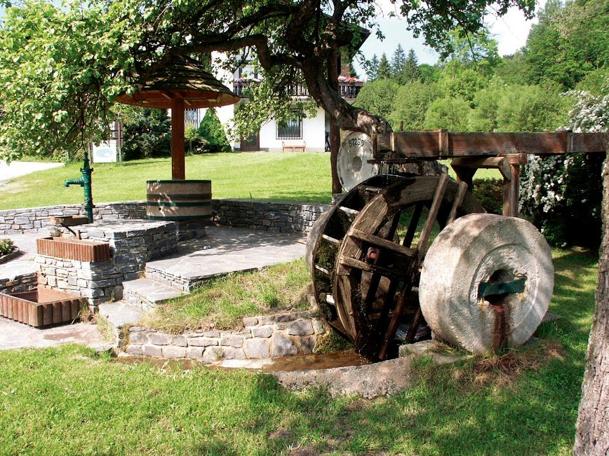 Old mill wheel with millstone, next to steps and a fountain, surrounded by meadow and trees.
