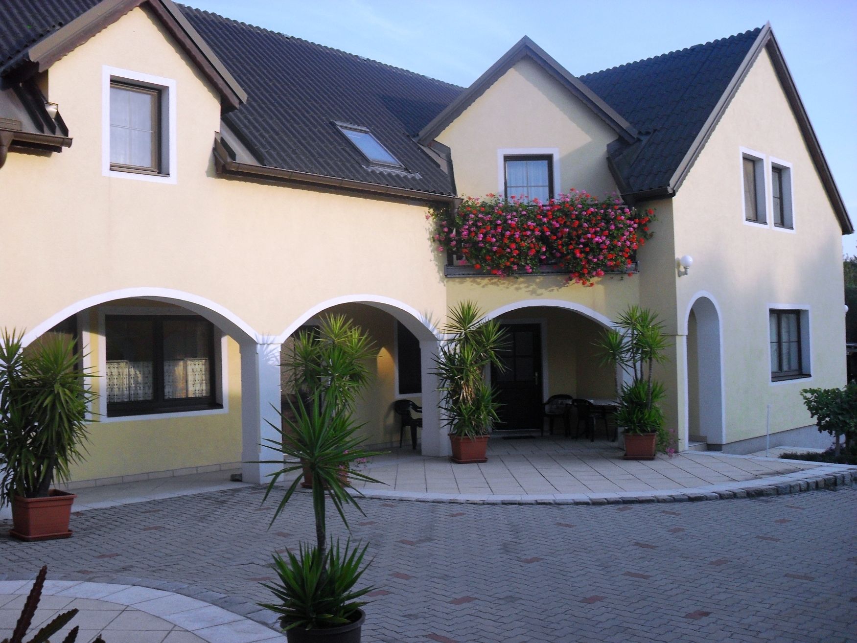 A yellow house with a dark roof, a balcony with flowers and plants in the courtyard.