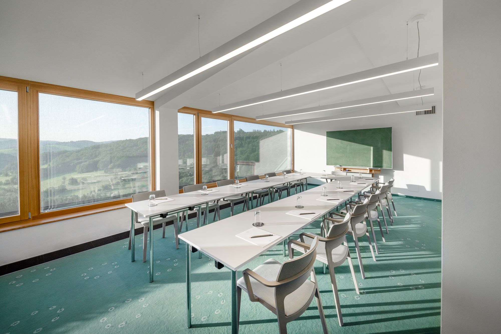 A bright seminar room with large windows offering a view of a green landscape. White tables and chairs are arranged in a U-shape.