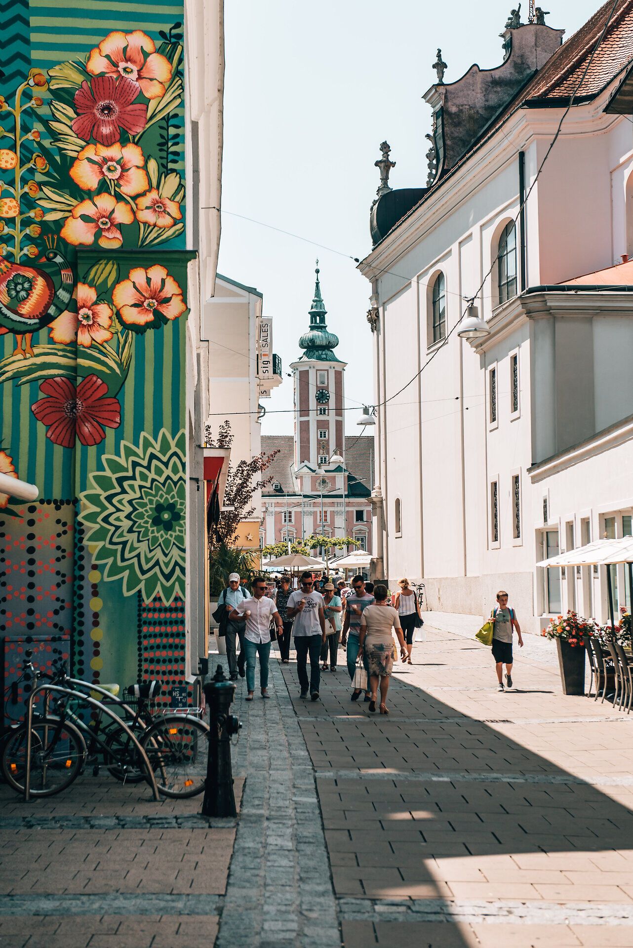 Die bunten Wandmalereien an den Gebäuden verleihen der Straße eine lebendige Atmosphäre, während die Menschen entspannt flanieren und die warmen Sonnenstrahlen genießen. Der Blick auf den markanten Kirchturm im Hintergrund lädt dazu ein, die charmante Stadt zu erkunden und die kulturellen Schätze zu entdecken.