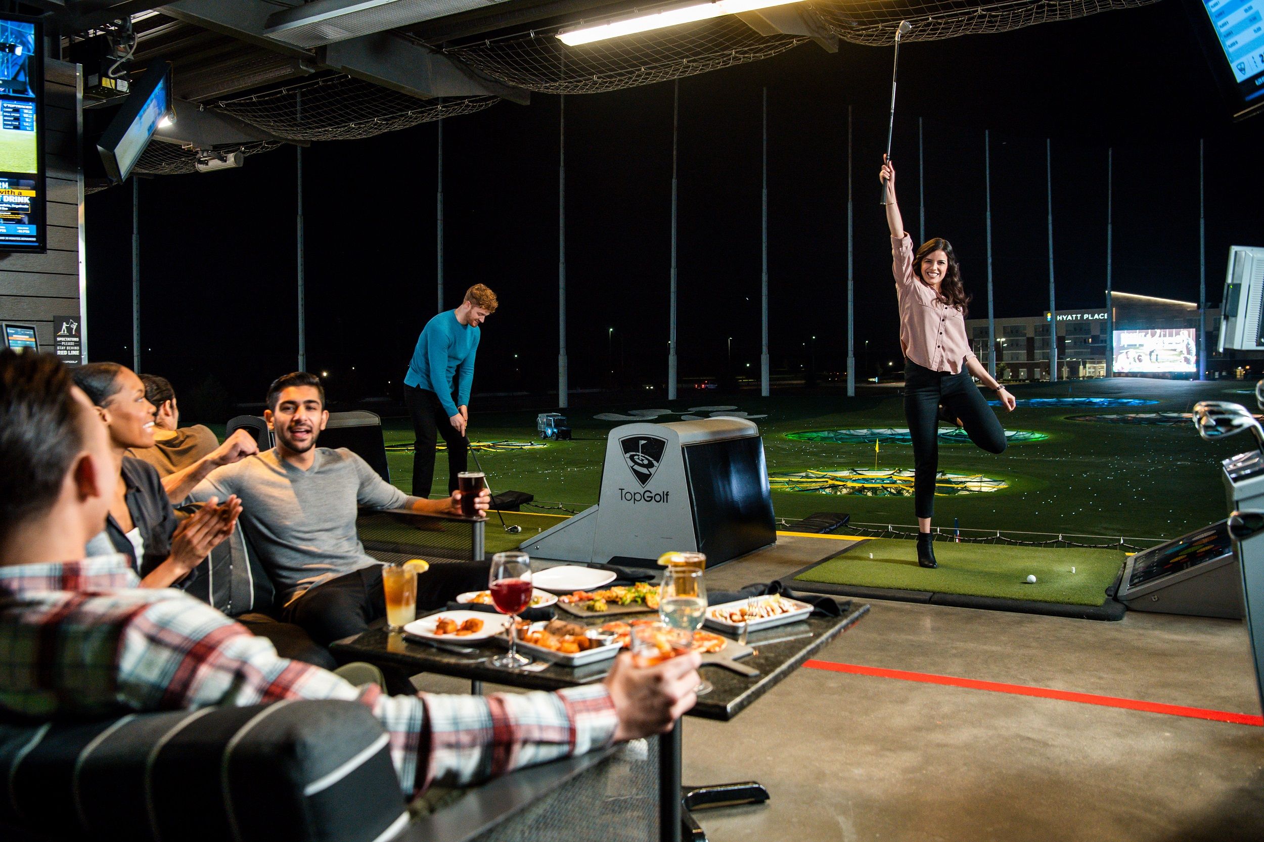 People play golf at TopGolf, a woman cheers while others eat and drink.