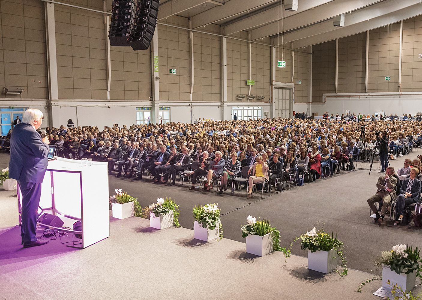 A speaker addresses a large audience in an exhibition hall.