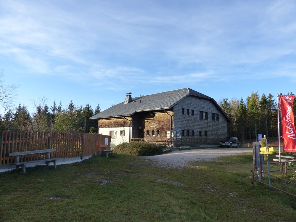 A rustic building with wooden walls and a gray roof, surrounded by trees. A red flag is on the right of the picture.