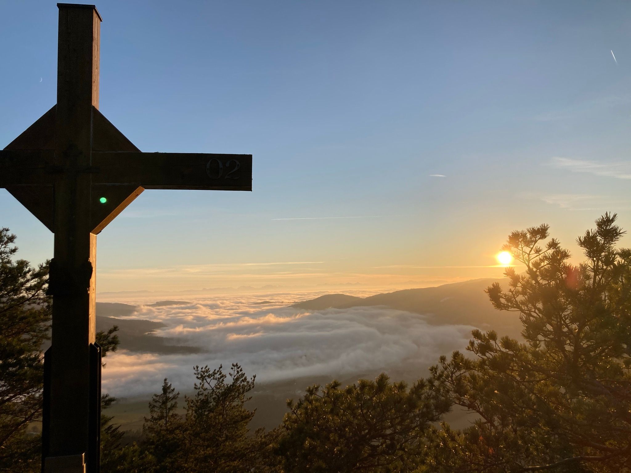 A summit cross on a mountain with a view of a sea of fog and a sunrise.