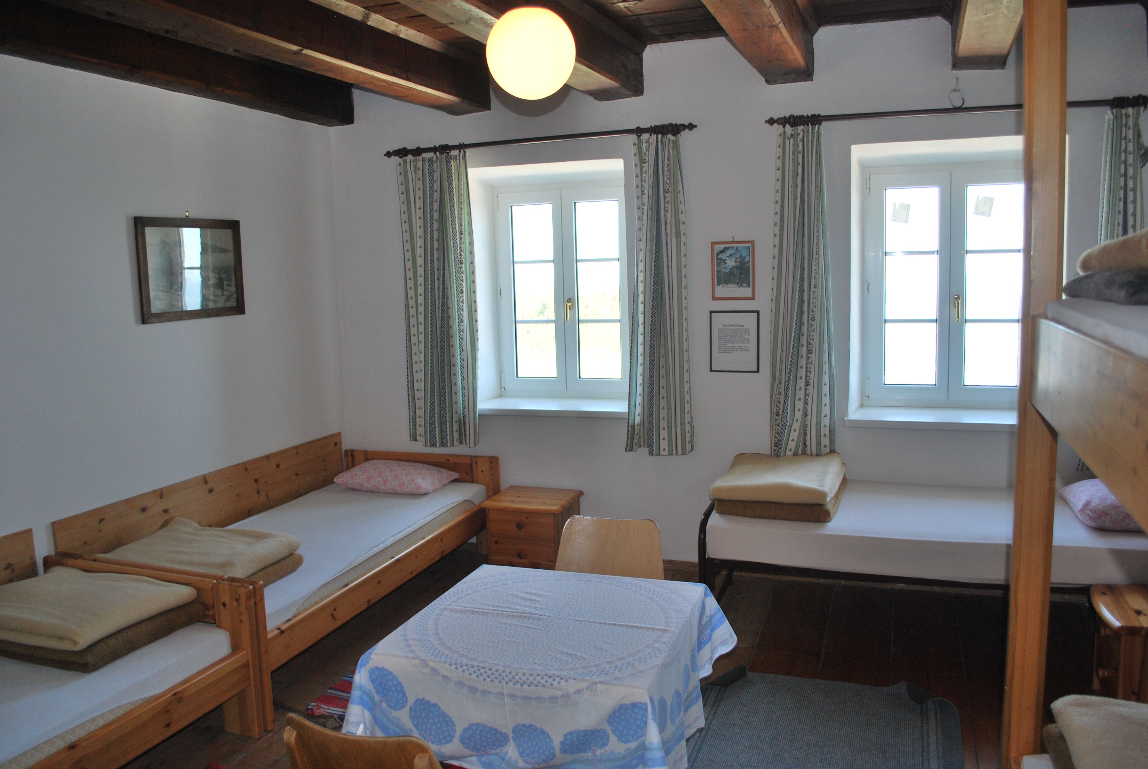 A simply furnished room with wooden furniture, two beds, a table and two windows with curtains.