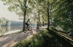 Zwei Personen, die man von hinten sieht, umgeben von Wald und Wasser, die mit ihren Rädern fahren