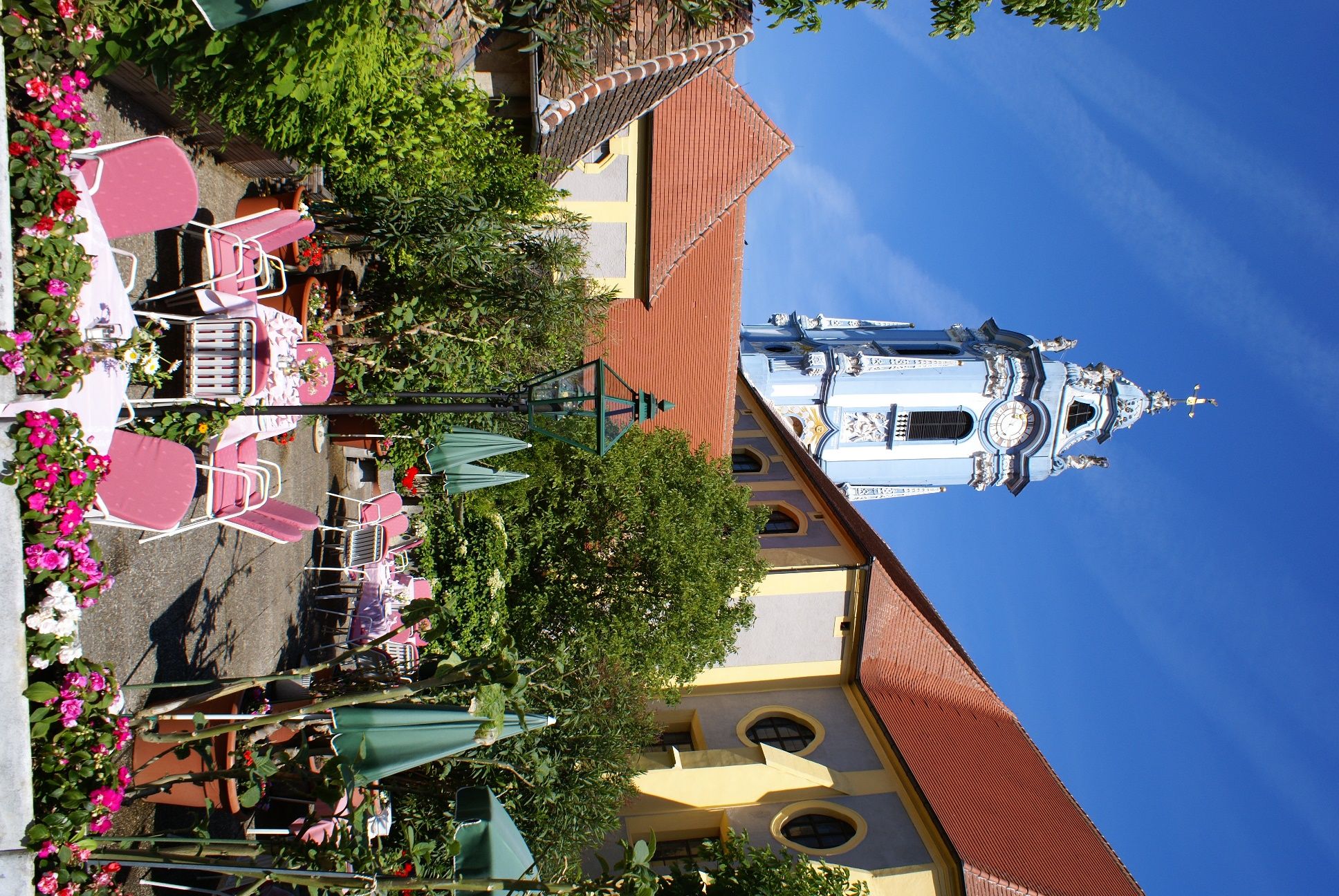 A flowering garden with pink chairs and tables in front of a historic building with a blue tower.