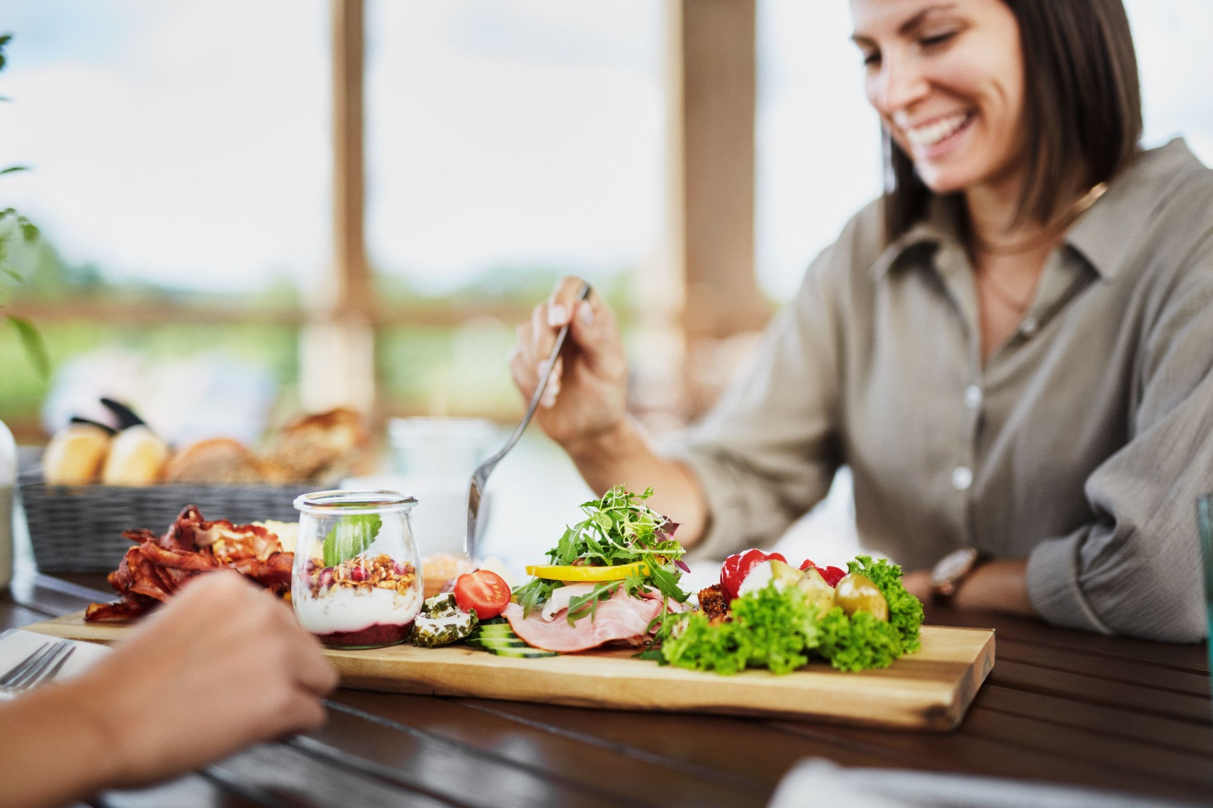 A woman eats breakfast with fresh ingredients on a wooden board on a terrace.