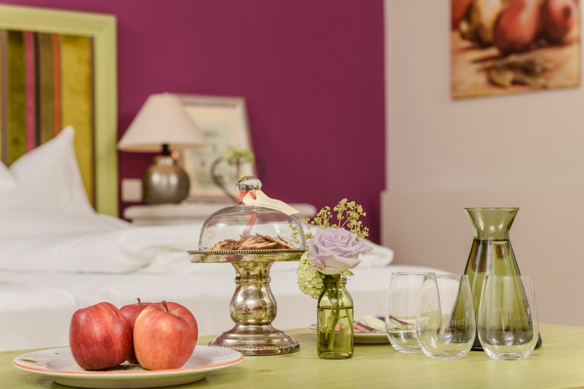 A stylishly furnished room with a table on which apples, cookies under a glass bell jar, a vase with a flower and glasses are placed.
