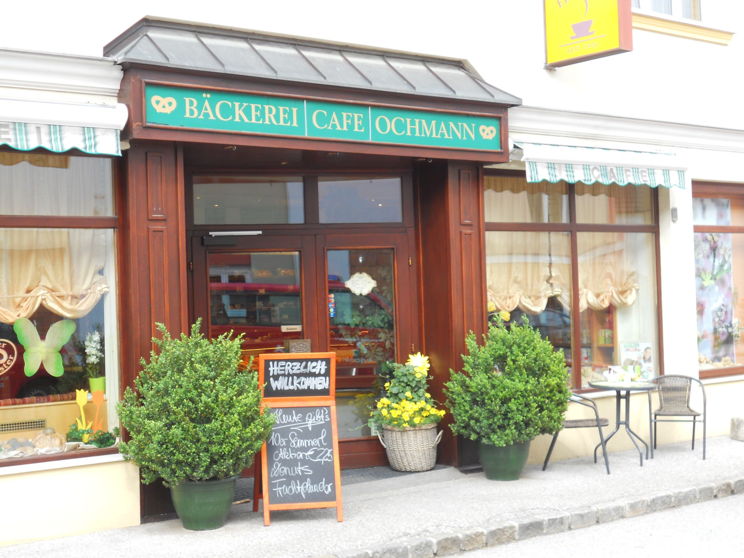 Entrance of a bakery, decorated with plants, a sign and a small table with chairs.