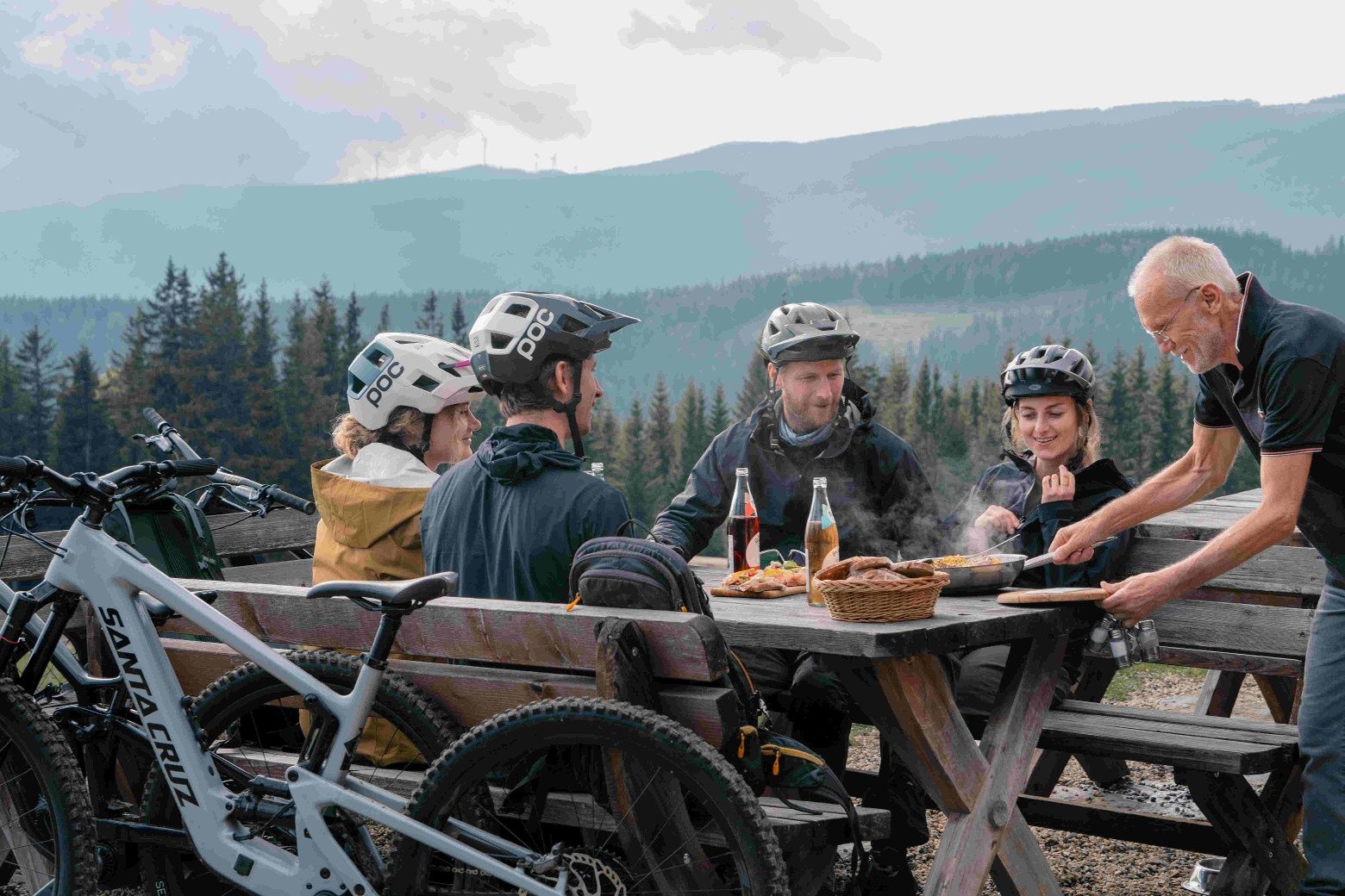 Group of mountain bikers sitting at a wooden table in the mountains enjoying a meal.