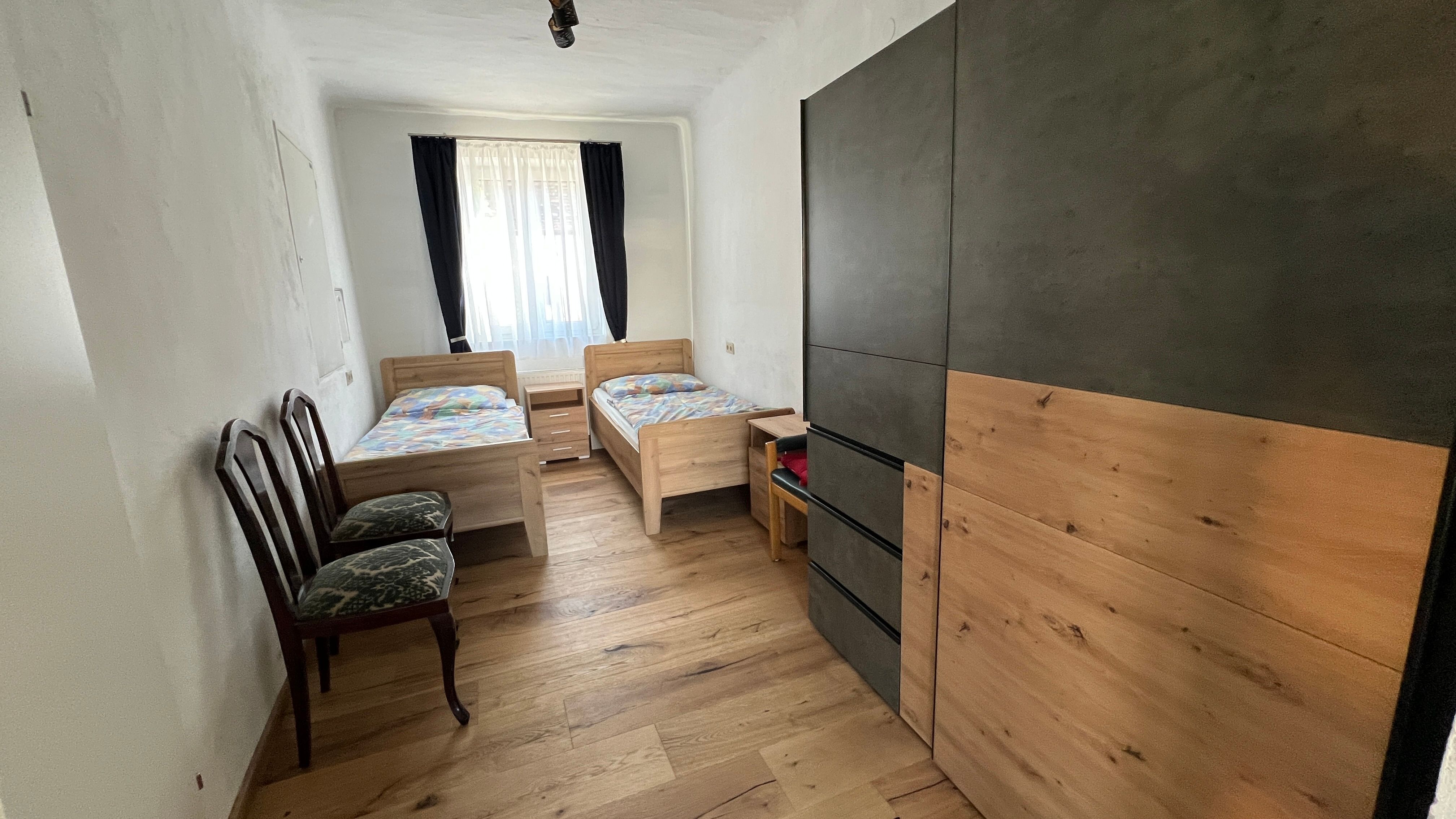 A small bedroom with two single beds, a wardrobe and two chairs. A window with curtains lets in light.
