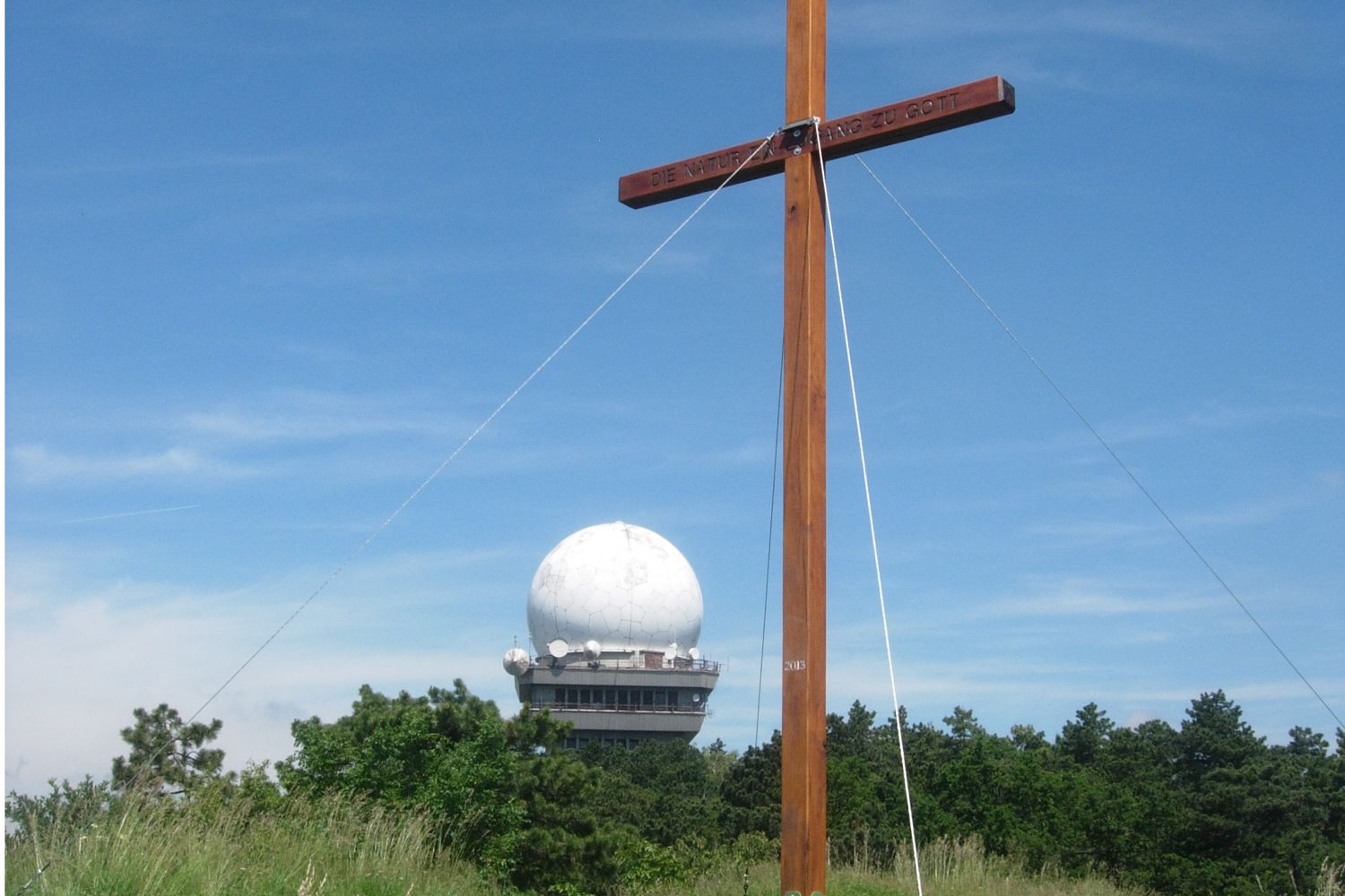 A wooden cross in front of a radar dome on the Buschberg, surrounded by trees and blue sky.