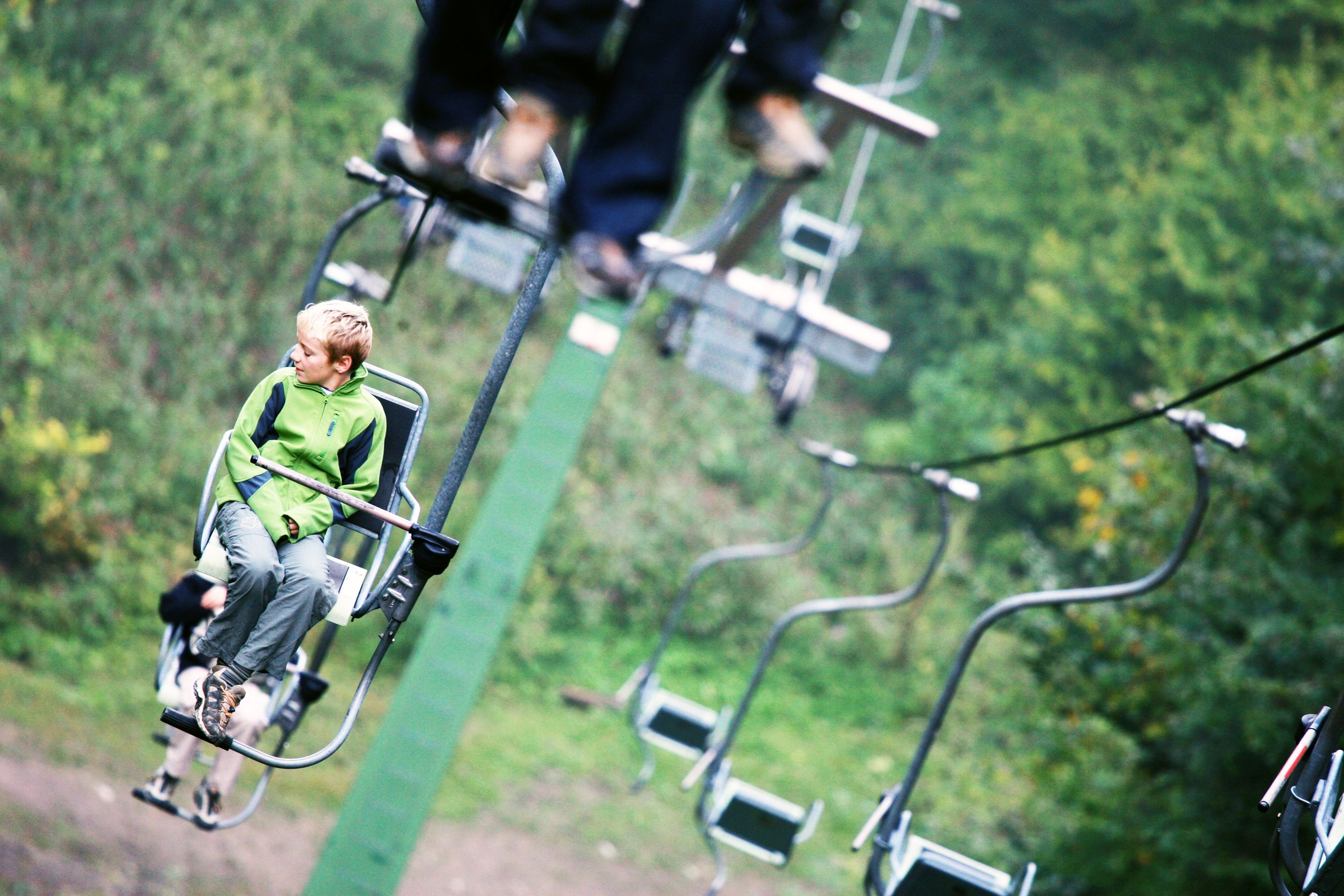 A boy in a green jacket sits on a chair lift, surrounded by green nature.