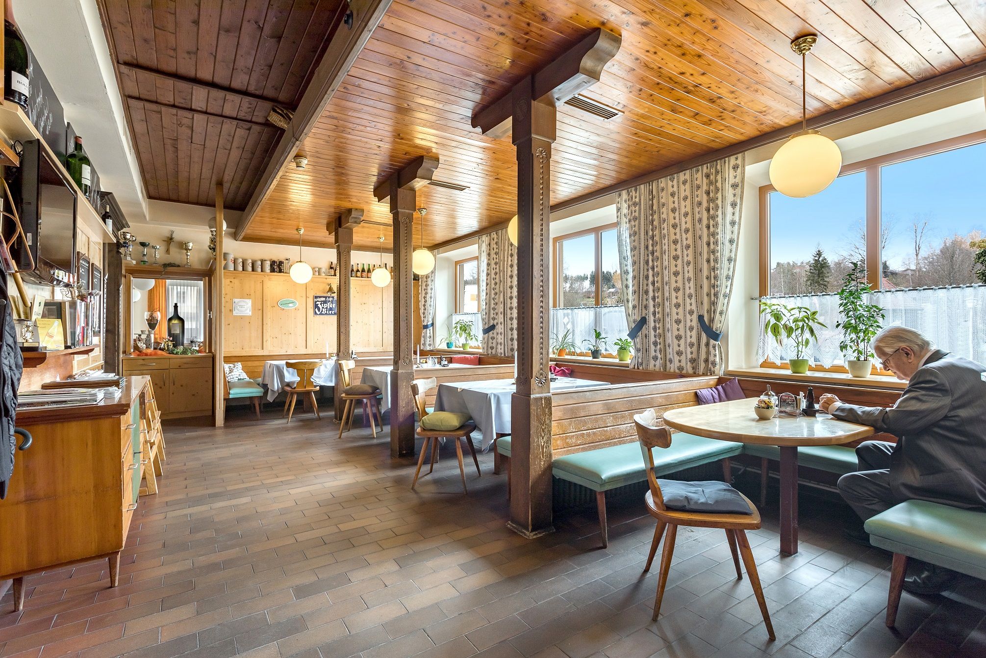 Cozy dining room with wooden furniture and large windows, a man is sitting at a table.