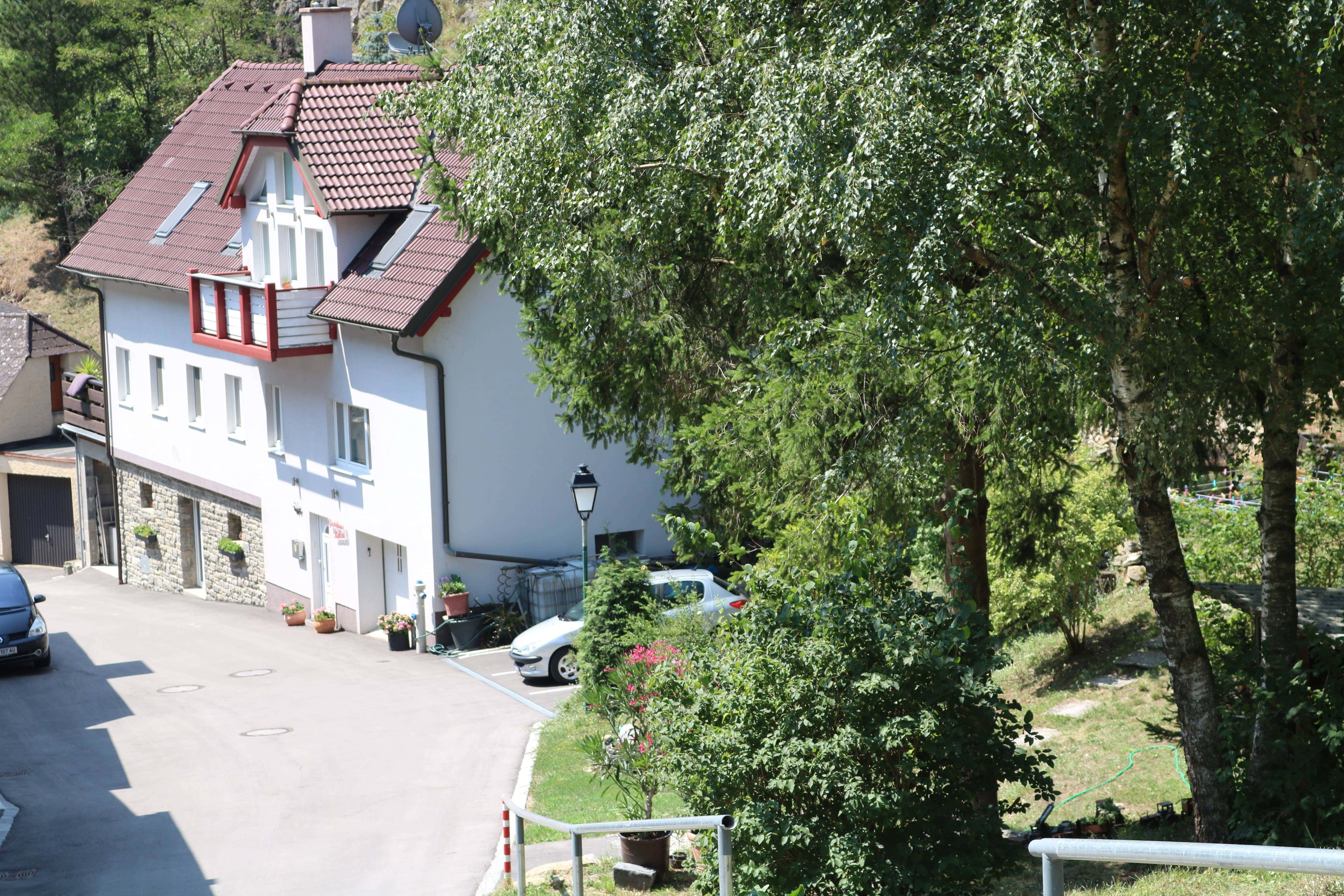Exterior view of a house with a red roof and white walls, surrounded by trees and a garden.