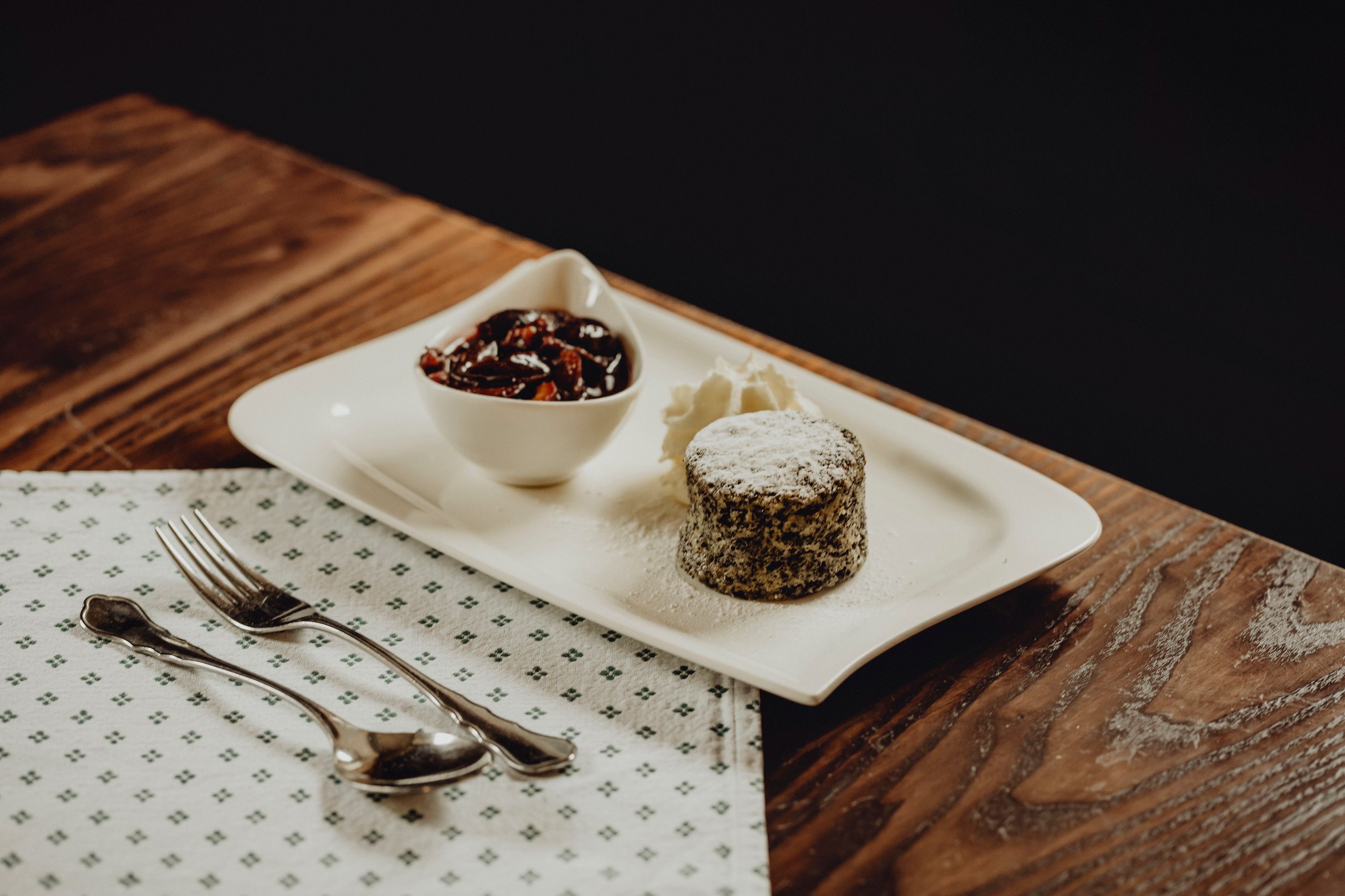 A poppy seed cake with powdered sugar, cream and a bowl of fruit on a white plate.
