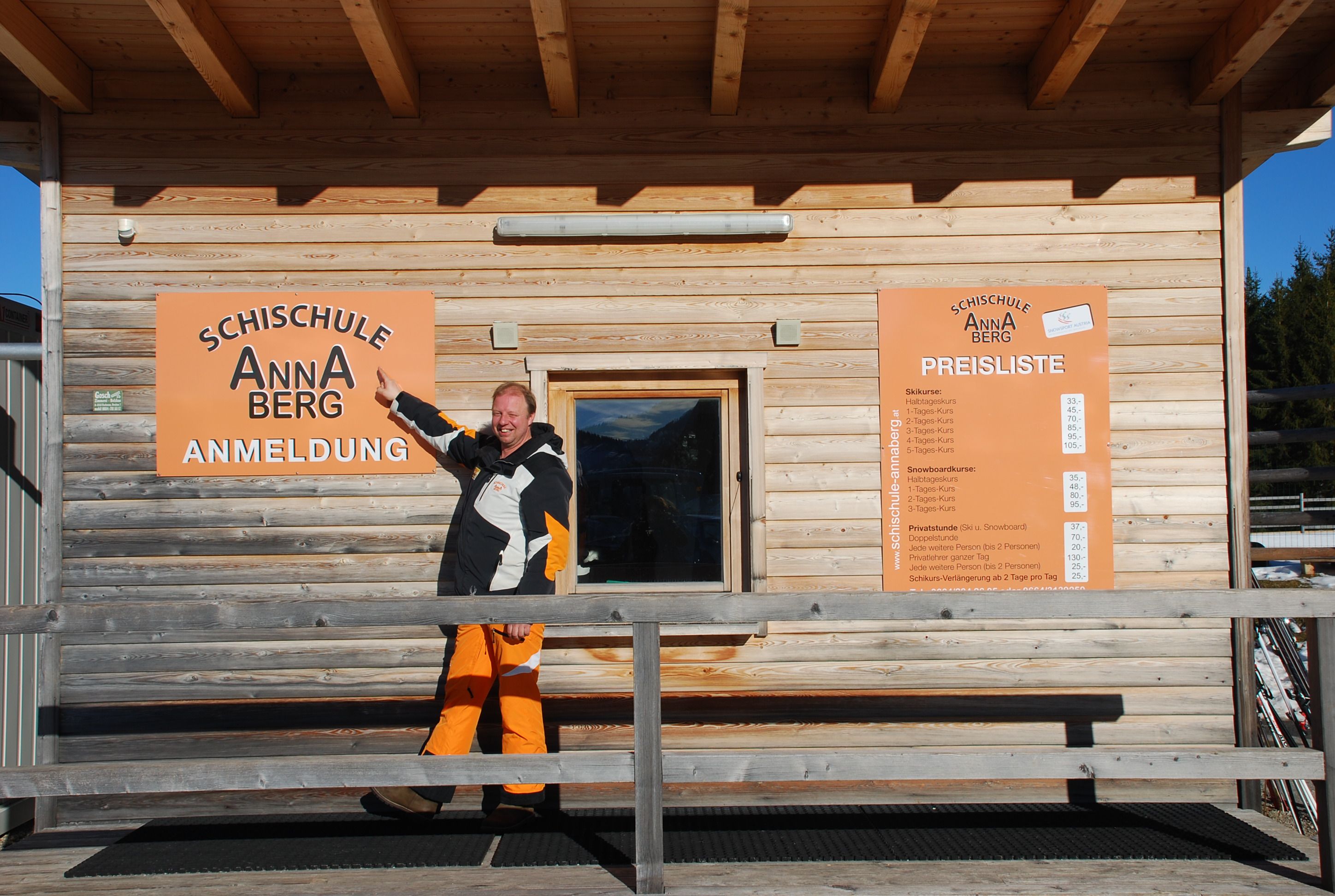 A man points to a sign for the Annaberg ski school in front of a wooden building.