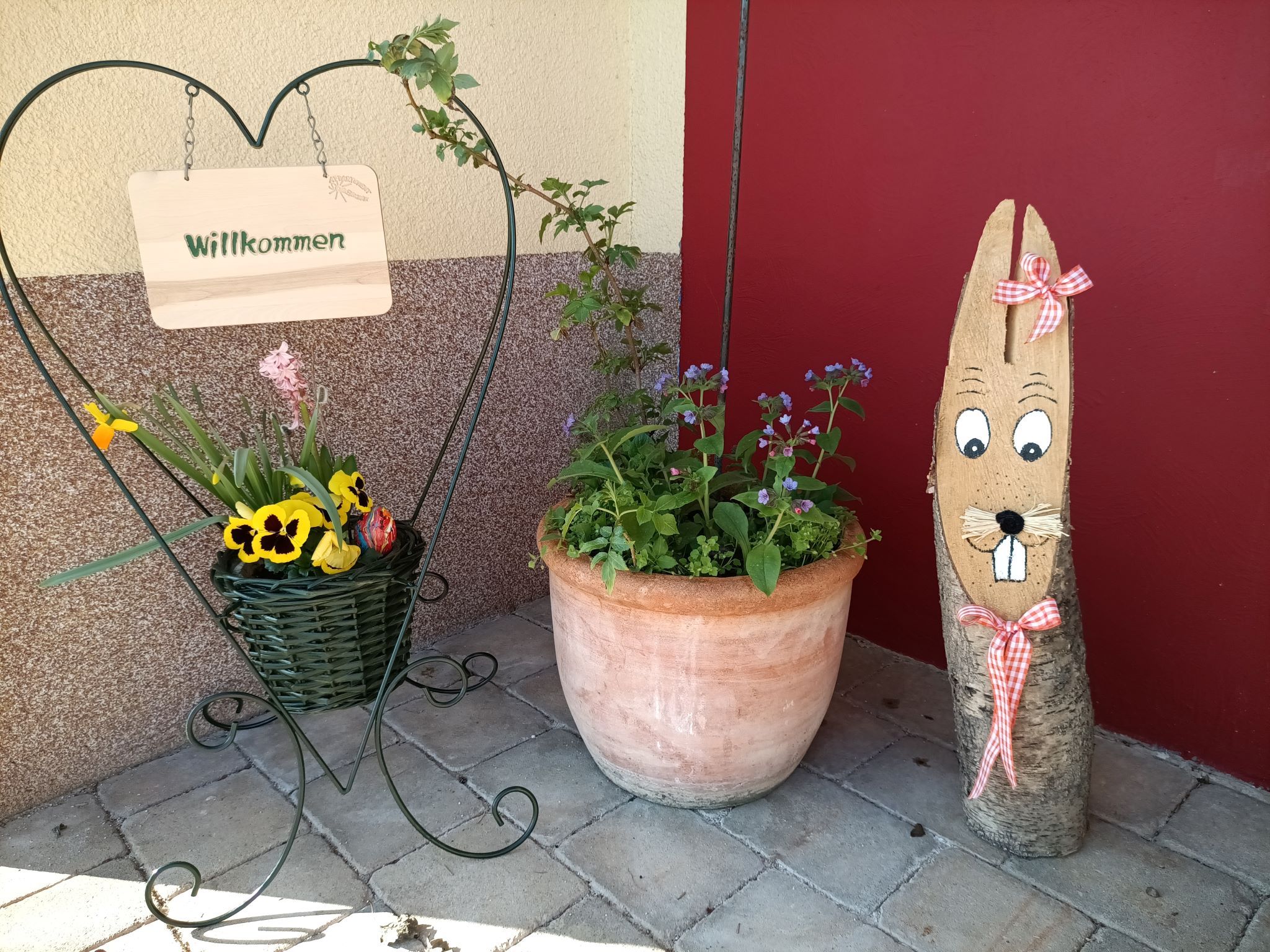 Welcome sign in the shape of a heart with flowers and wooden figure in front of a red wall.