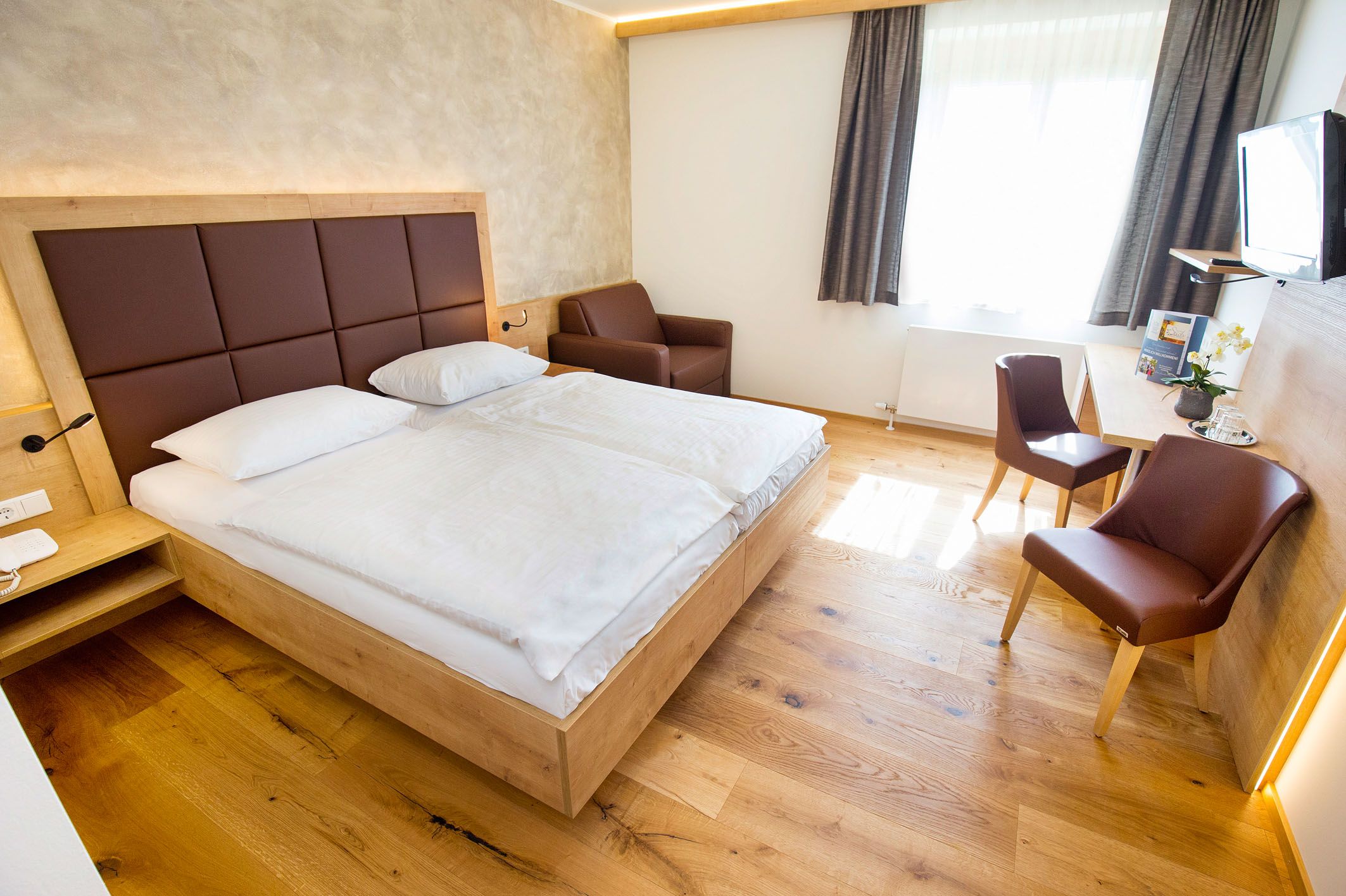 Modernly furnished hotel room with double bed, desk, chairs and an armchair.