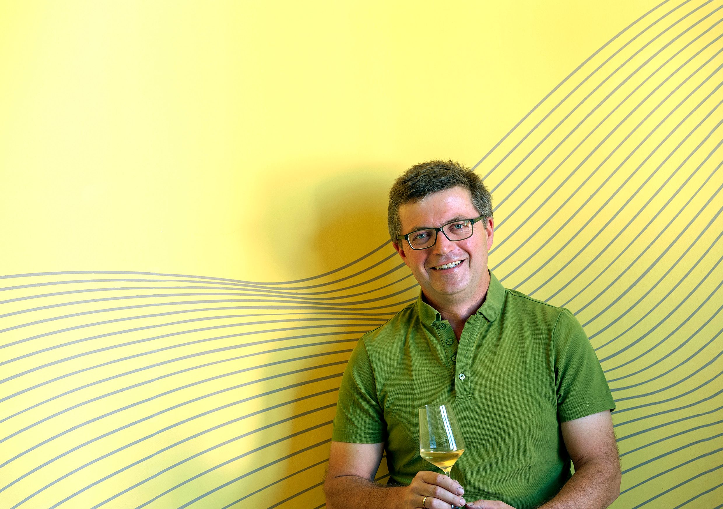Man with glasses and green shirt holds a wine glass in front of a yellow background with a line pattern.