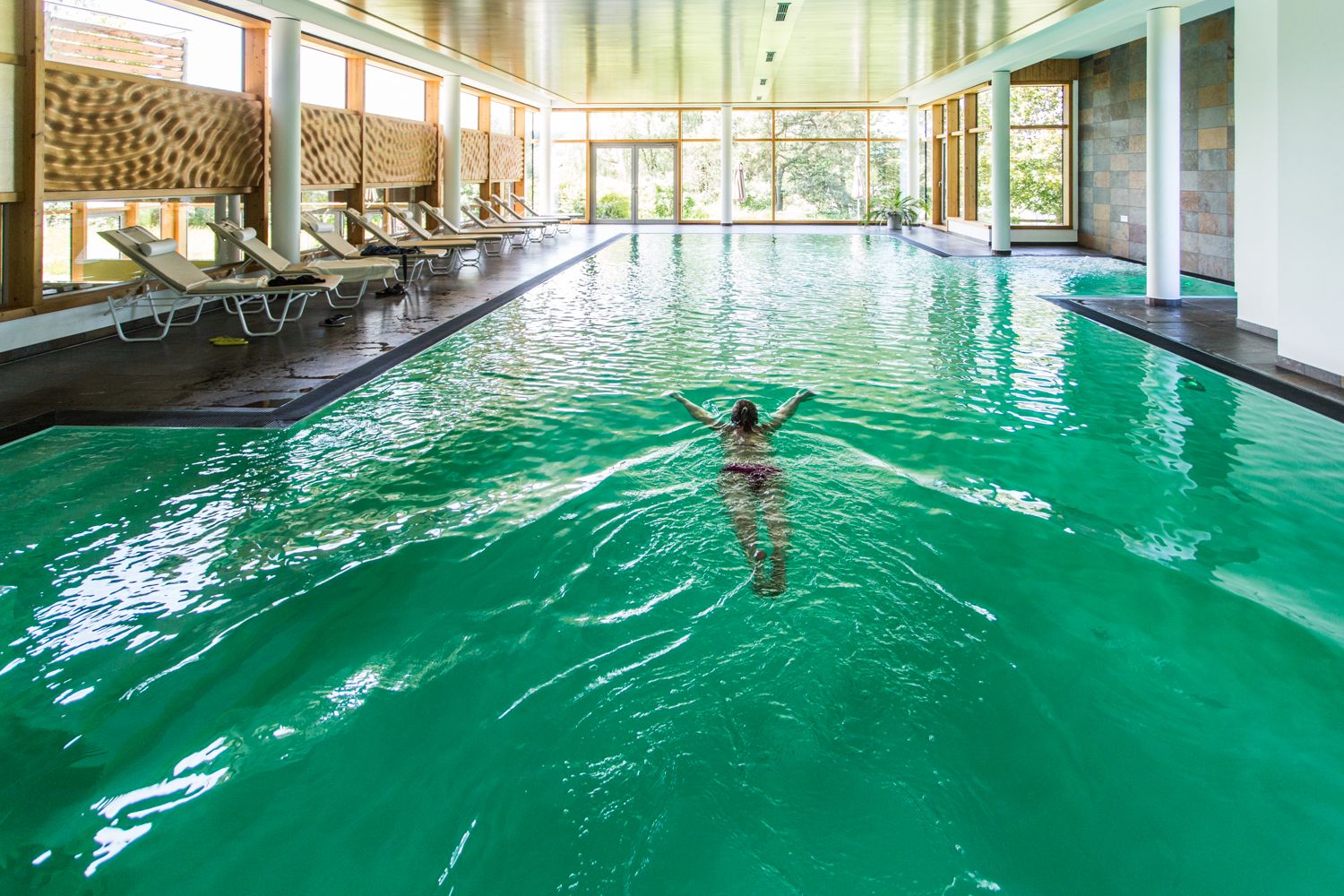 Person swimming in an indoor pool with green water and deckchairs at the edge.