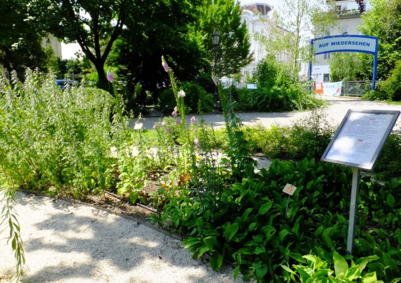 A well-tended garden with plants and a sign, in the background a gate with the inscription 'Auf Wiedersehen'.