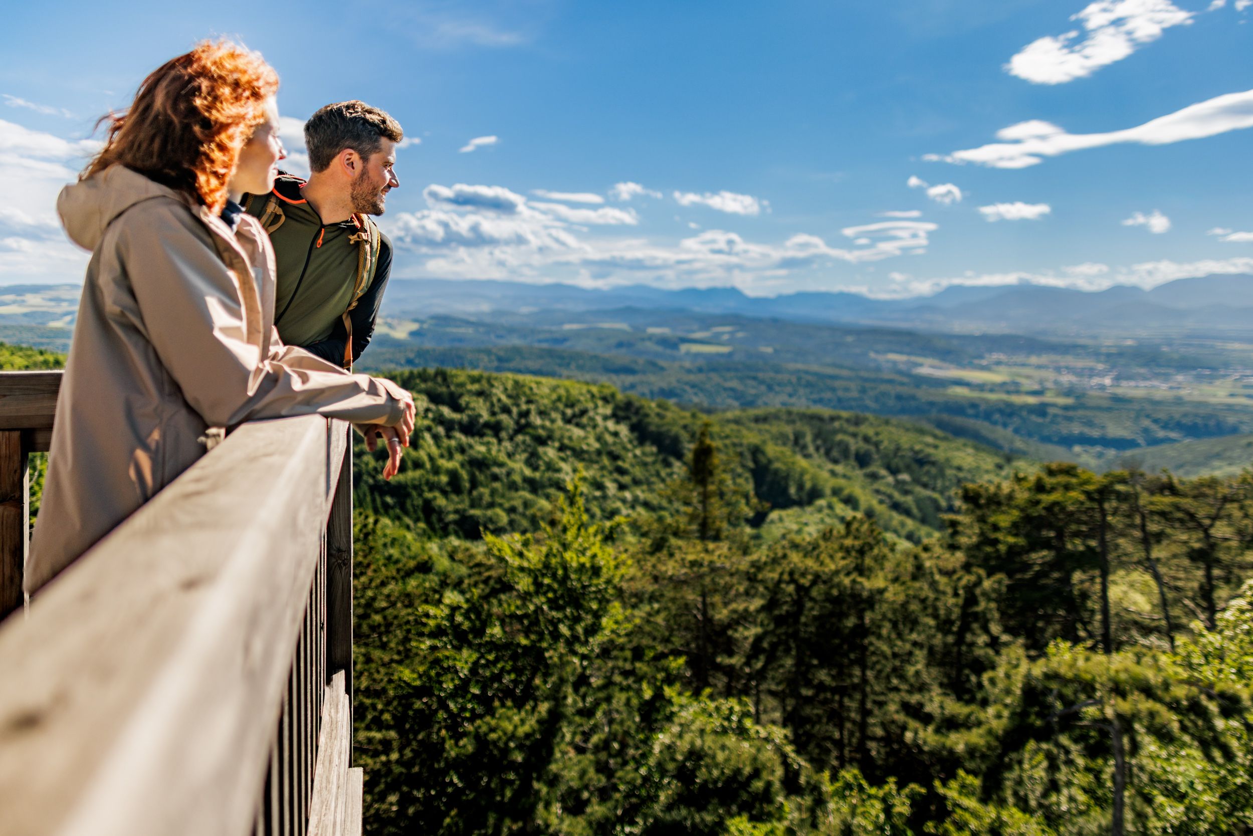 Two people stand on the platform of the Lanzenkirchen/Wiesen observation tower and look out over a wooded landscape with mountains in the background.