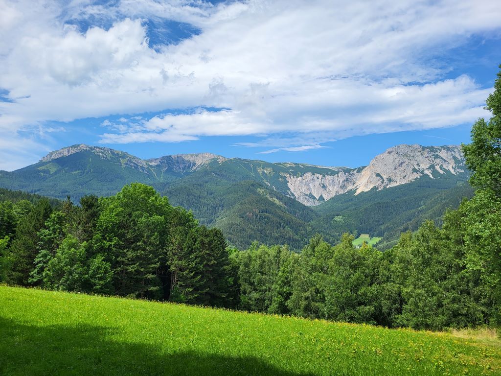 Mountain landscape with green meadows and wooded hills under a blue sky.