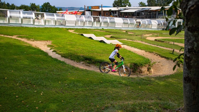 A child with an orange helmet rides a green bike through a mini bike park with hills and bends.