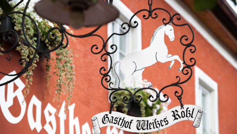 Metal sign with a white horse and the words 'Gasthof Weißes Rößl' in front of a red building.