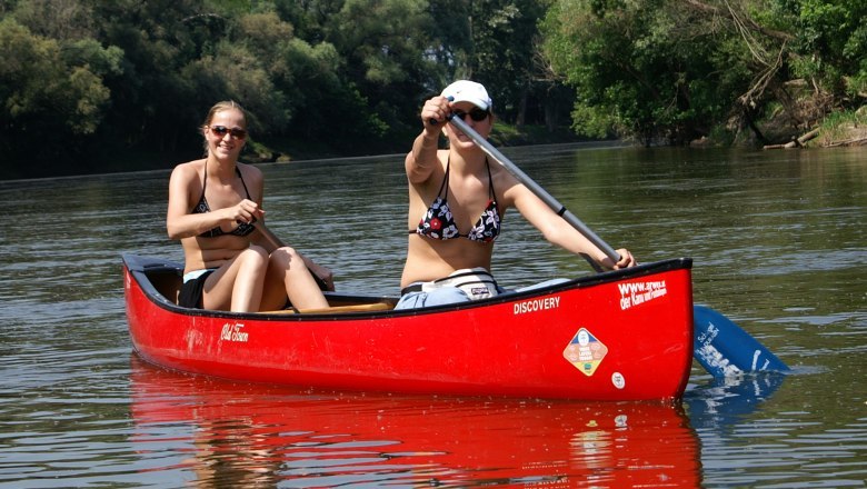Two women paddle in a red canoe on a river, surrounded by green nature.
