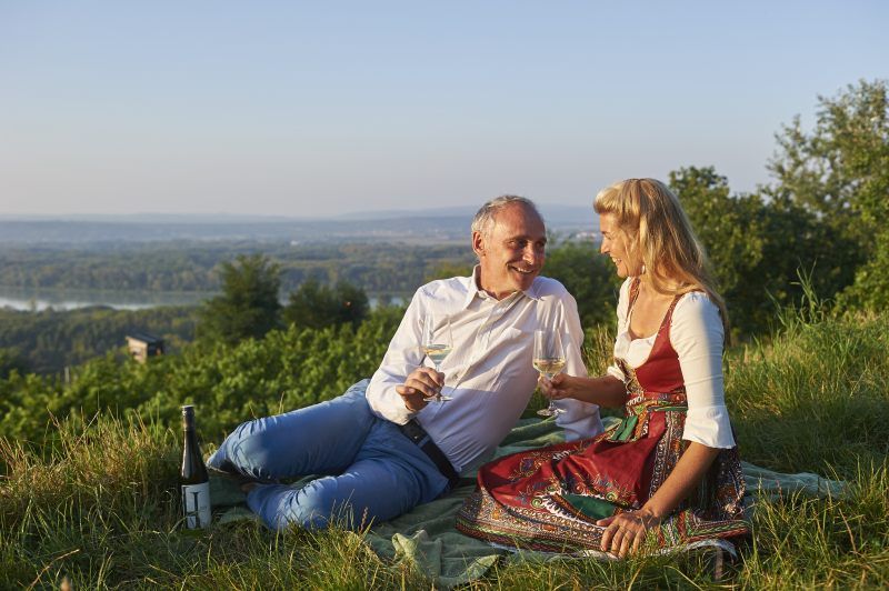 A couple sits on a meadow and drinks wine with a view of a landscape.