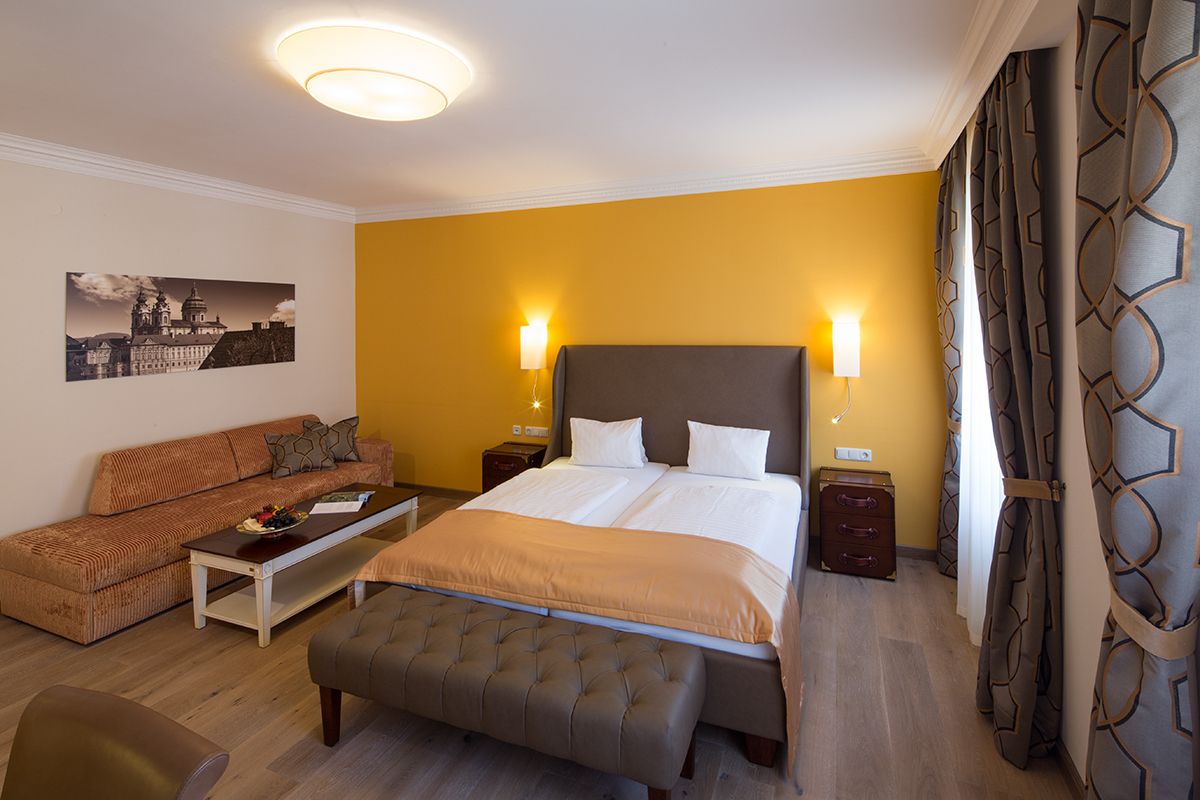 Hotel room with double bed, yellow wall, sofa, table and curtains.
