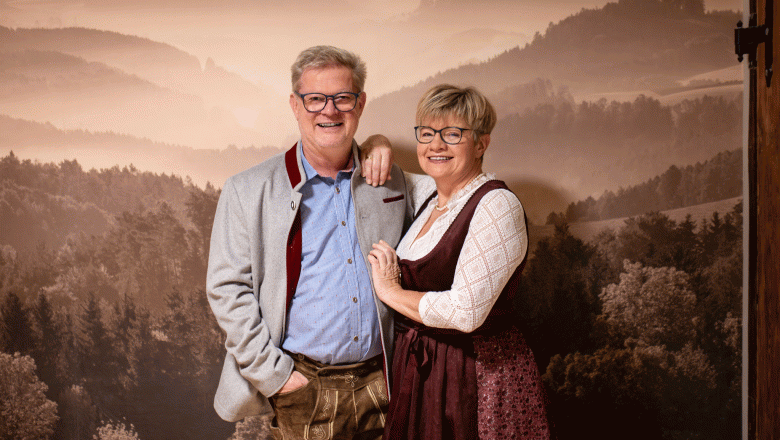 A couple in traditional Bavarian dress stands in front of a background with a forest landscape.