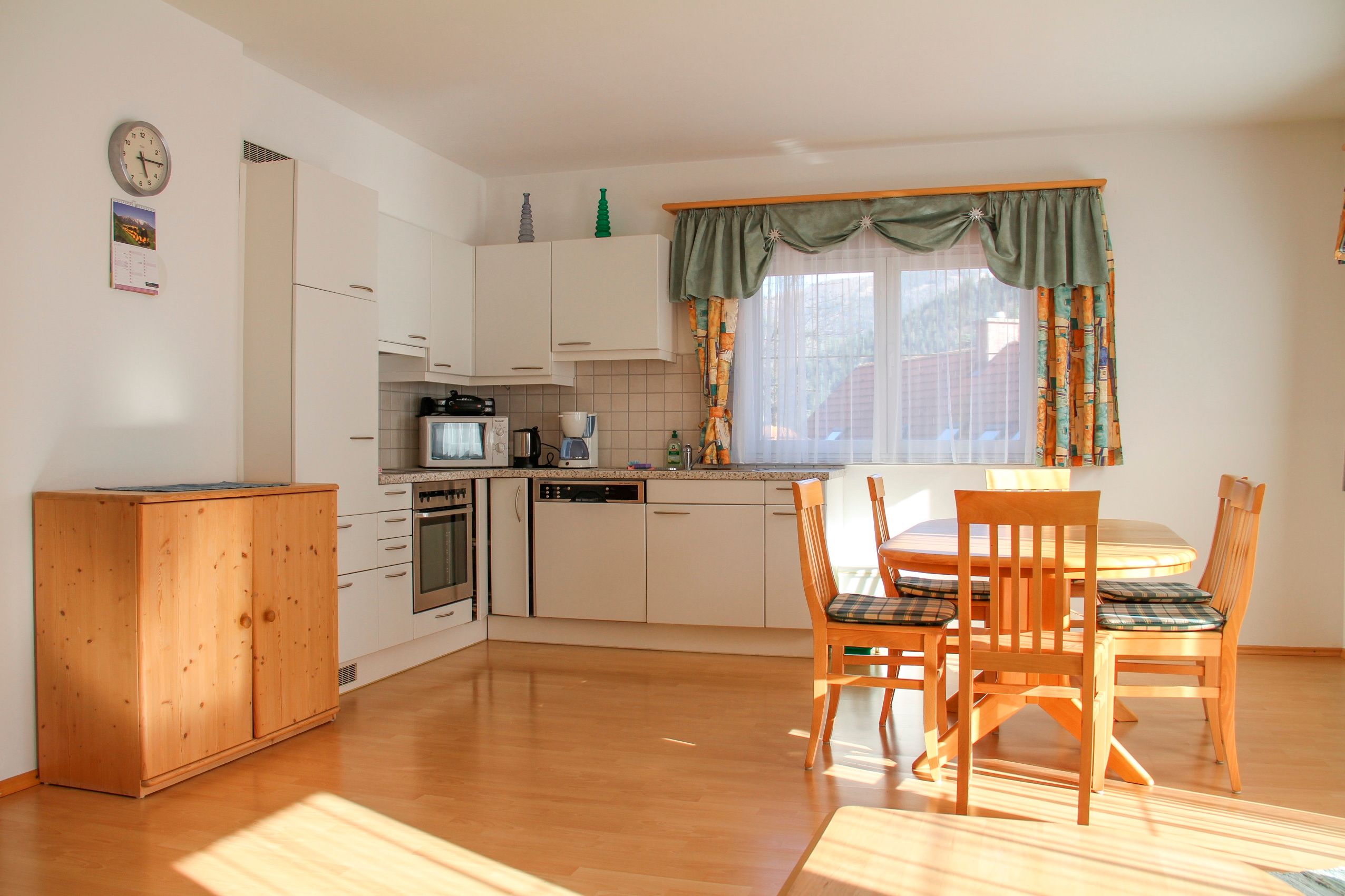 Bright kitchen with dining table and chairs, white cupboards, wooden floor, windows with curtains.