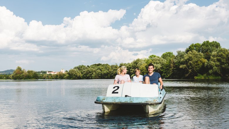 Three people on a pedal boat on a lake with trees in the background.