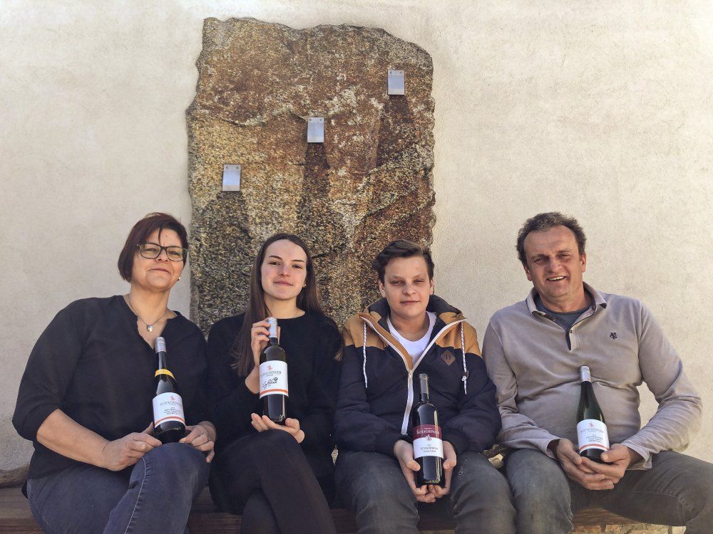 Four people sit in front of a stone wall holding bottles of wine.