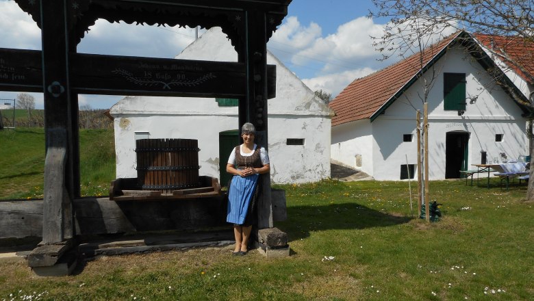A woman in traditional dress stands next to an old wine press in front of a historic building with white walls and red roofs.