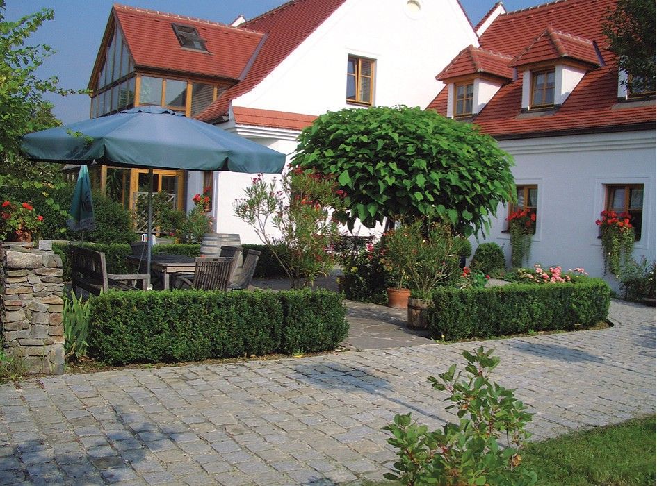 A well-kept garden with paving stones, a large parasol, wooden furniture and a white building with red roofs in the background.