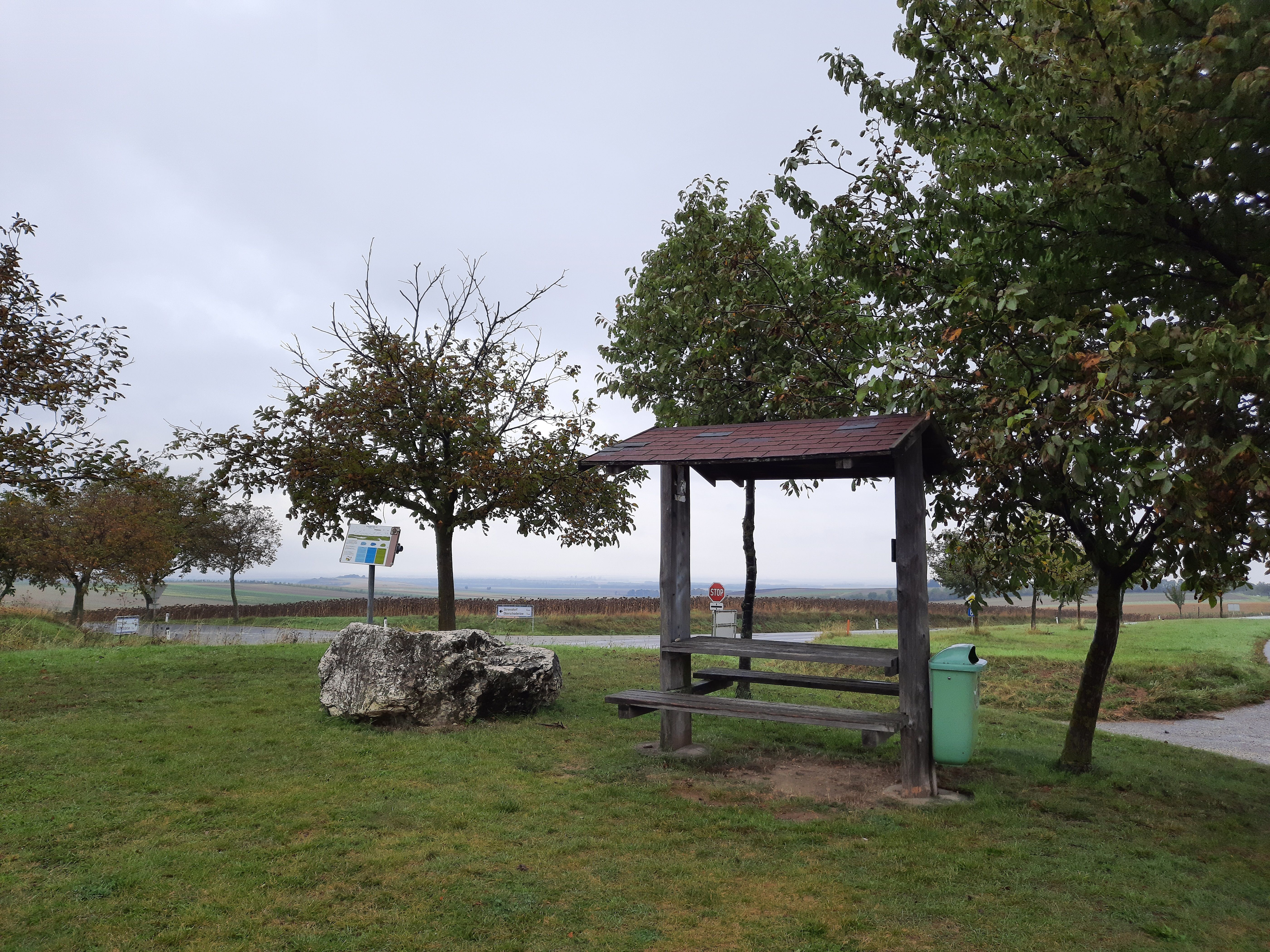 A rest area with wooden benches and a table, surrounded by trees and a meadow, under a cloudy sky.