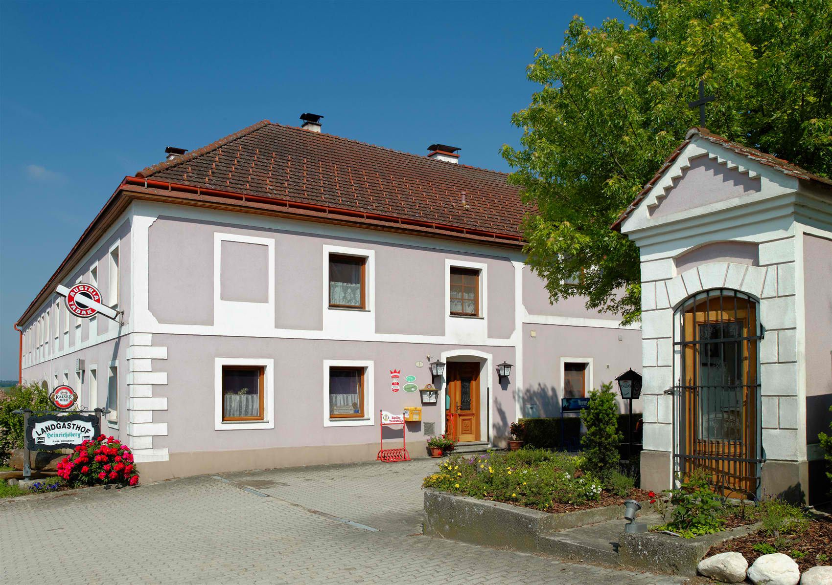 A traditional country inn with a gray façade and red roof tiles, surrounded by flowers and trees.