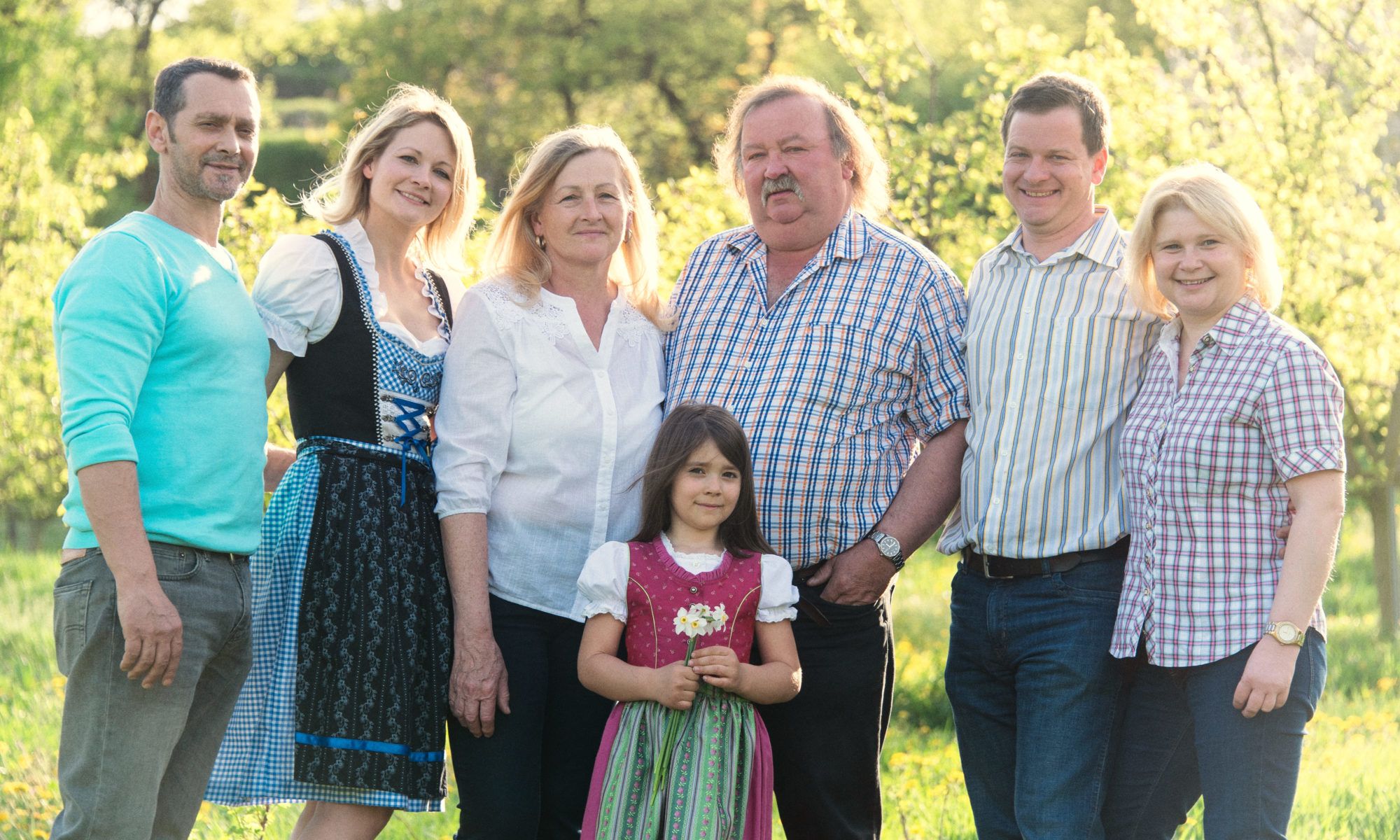 A smiling family stands in a sunny garden, some wearing traditional clothing.