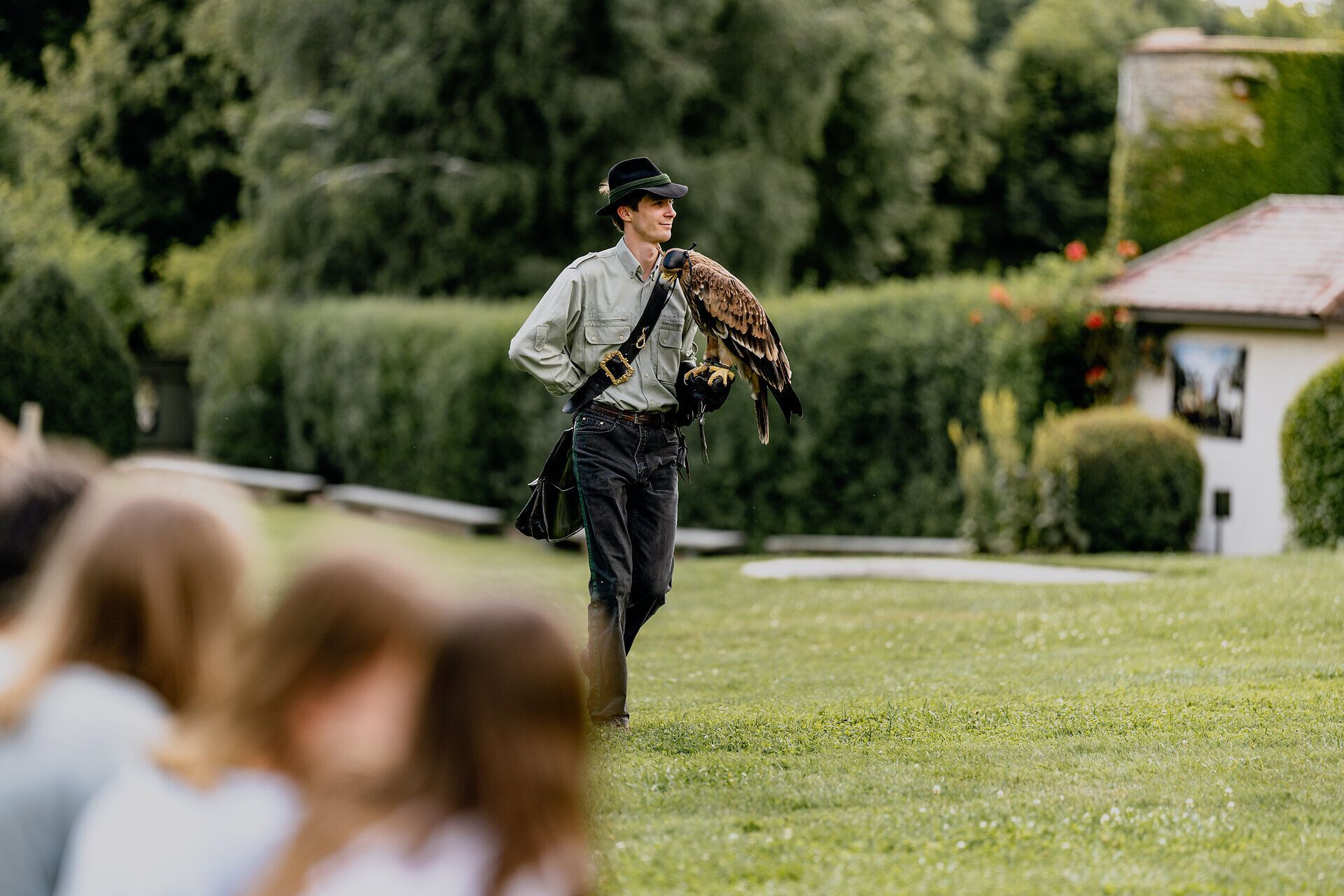 A falconer with a bird of prey on his arm stands on a meadow in front of a group of spectators.