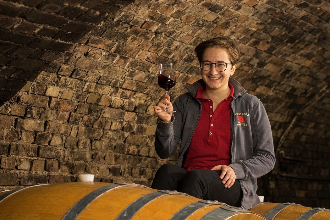 A person sits on a wine barrel in a wine cellar and holds a glass of red wine in his hand.