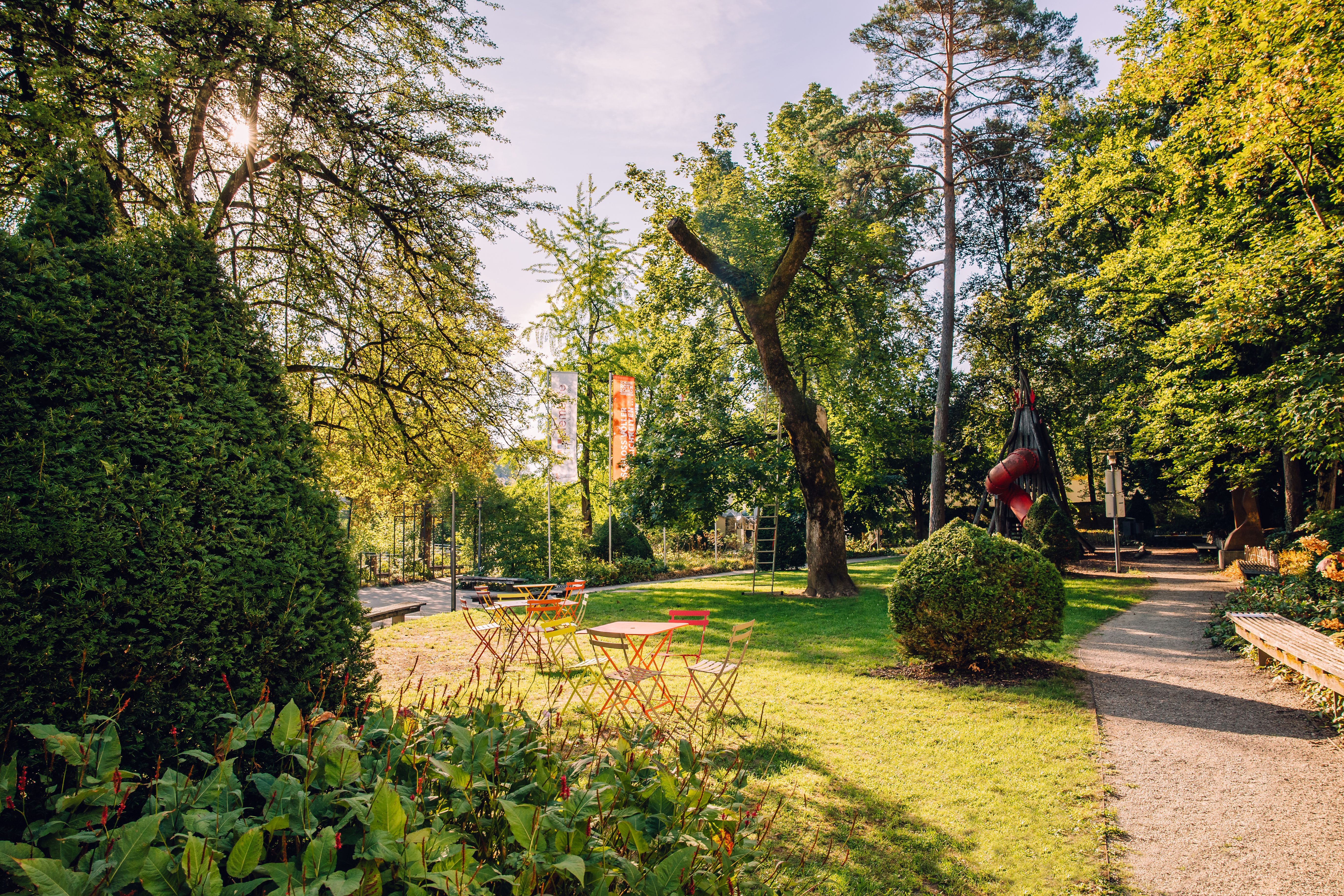 A sunny park at Schloss Rothschild in Waidhofen an der Ybbs, featuring colorful seating areas, old trees, and walking paths.